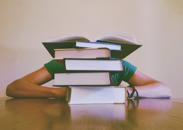 A person sits with their head buried behind a stack of books on a desk.